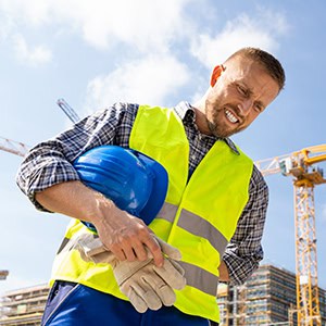 A worker in a yellow safety vest holds a blue hard hat at a construction site under a clear sky. - Leep Tescher Helfman and Zanze